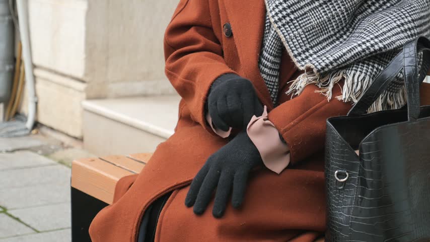 Person wearing long brown coat and plaid scarf sits on wooden bench outdoors and adjusts black gloves on hands while waiting in cold weather scene.