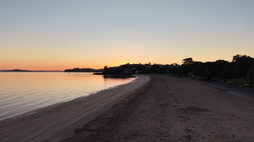 Aerial drone flight along calm beach shoreline at sunrise, eastern bays, Tamaki Drive, Auckland, New Zealand