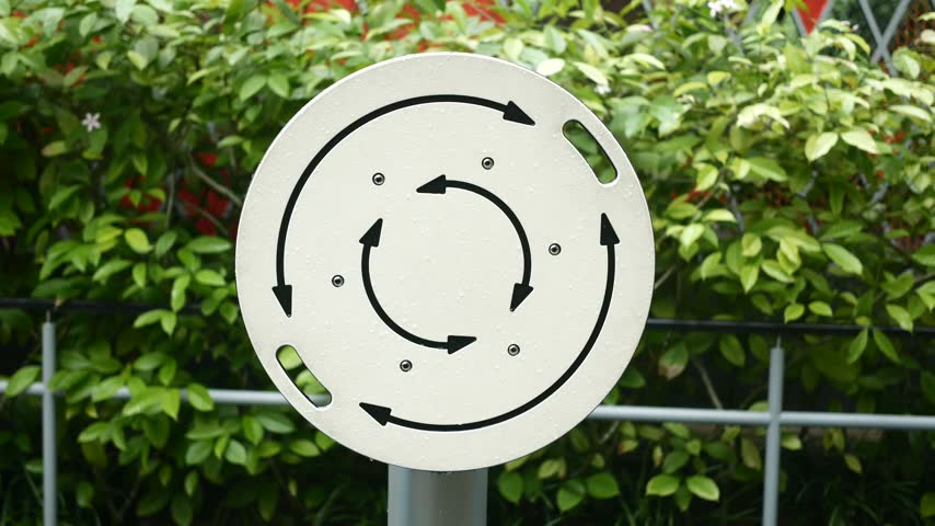 Person spins white circular exercise wheel with black arrow designs in public garden setting. Outdoor fitness station equipment rotates smoothly during bright daylight.