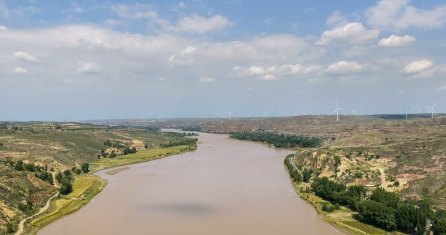 Aerial time-lapse of the muddy Yellow River flowing past loess plateau with wind turbines on distant ridges under blue sky, Hohhot city, Inner Mongolia, China.