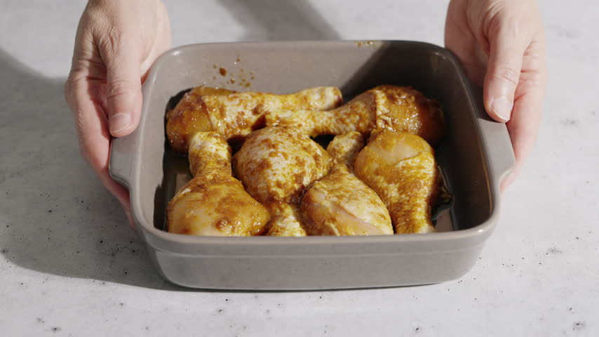 Hands Hold A Baking Tray Filled With Marinated Chicken Drumsticks.