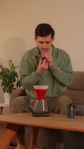 A close-up shot of a young man sitting on a couch and carefully pouring freshly ground coffee beans from a manual grinder cup into a red V60 dripper with a paper filter, set up over a glass server