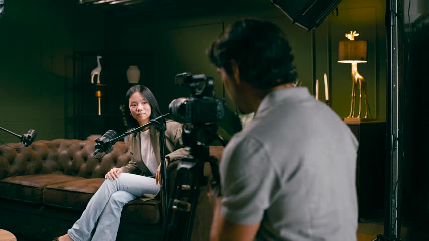 Young Asian podcaster sitting on a leather sofa during a live streaming interview. Professional cameraman using a camera to record the episode
