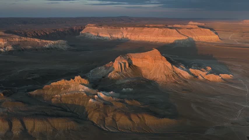 A gentle forward aerial passes over a serrated foreground ridge toward a sunlit Airakty butte, with dark gullies and a broad distant tableland providing depth.