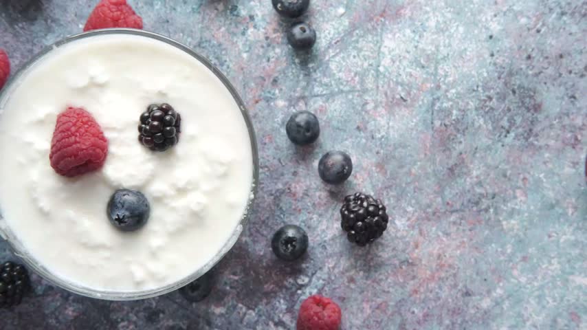 Top down close up of creamy white yogurt served in glass bowl decorated with fresh raspberries blueberries and blackberries scattered on rustic textured blue background healthy snack.