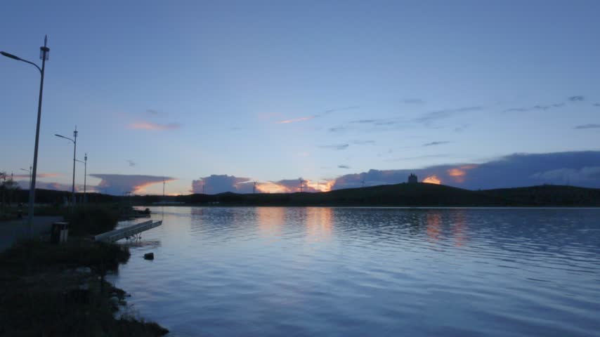Panoramic view of sunset silhouettes of orange light on the shore of a blue-toned twilight lake.
