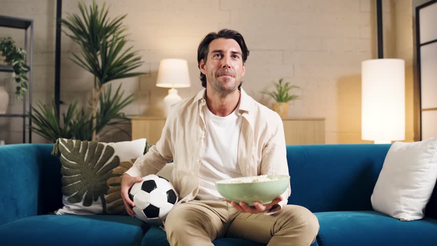 Passionate young man sitting on a couch, holding a soccer ball and popcorn. He watches television with excitement, celebrating his team