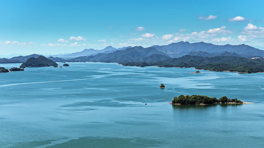 High angle shot of clear turquoise lake waters dotted with green islets, framed by distant mountain ranges under sunny blue skies.
