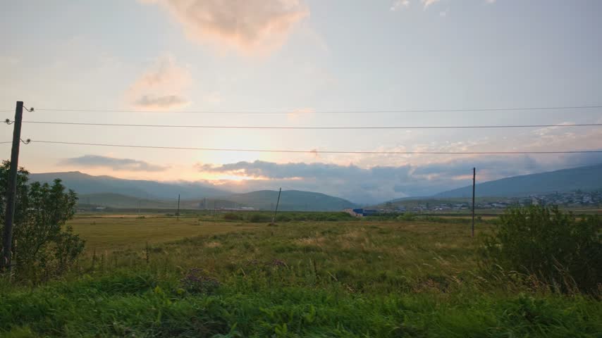 Sunset over green fields with distant mountains. Tall grasses sway in breeze. Wooden pole and power lines cross frame. Clouds glow in soft orange hues. Landscape peaceful and natural