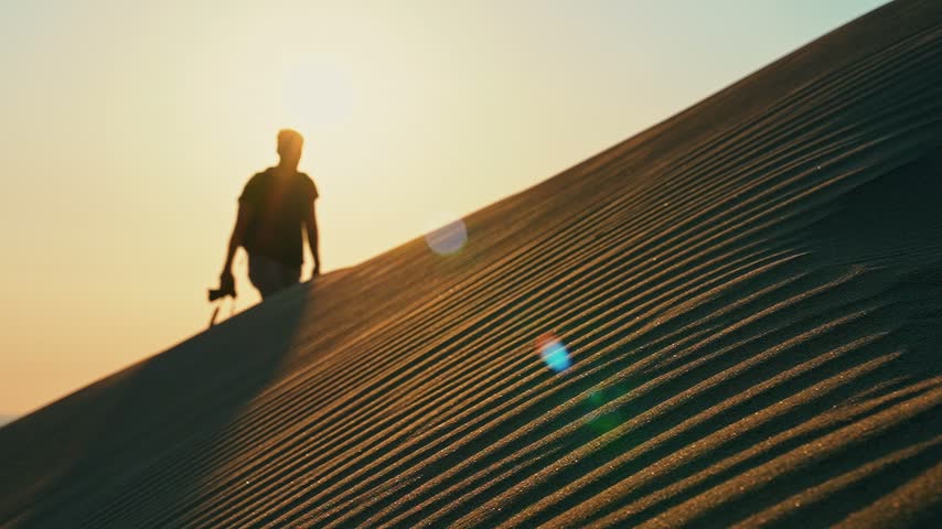 Desert sand dunes at sunset reveal a photographer walking in slow motion as golden sunlight illuminates the landscape and sand grains drift in the wind.