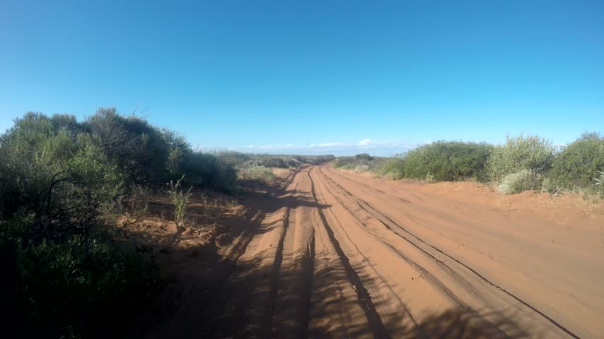 A red dirt road with parallel tire tracks runs through dry scrub and open Australian countryside.