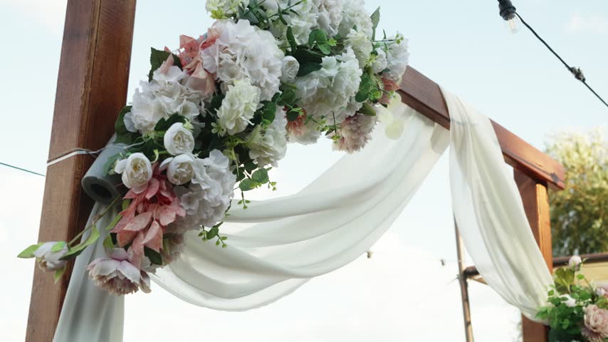 Wooden wedding arch decorated with white and pink flowers and flowing fabric against the sky