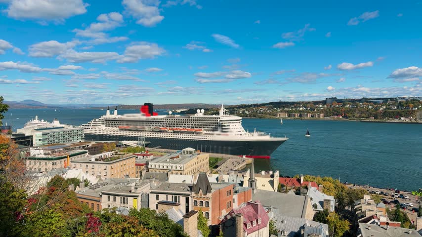 Wide shot of a large transatlantic ocean liner docked at the port of Quebec City in Canada under a blue sky with fluffy white clouds. Majestic cruise ship stands near the historic lower town buildings next to the St Lawrence River. Scenic view captures the large passenger vessel during a beautiful autumn day. Elevated perspective shows this iconic watercraft surrounded by urban architecture and harbor waters.