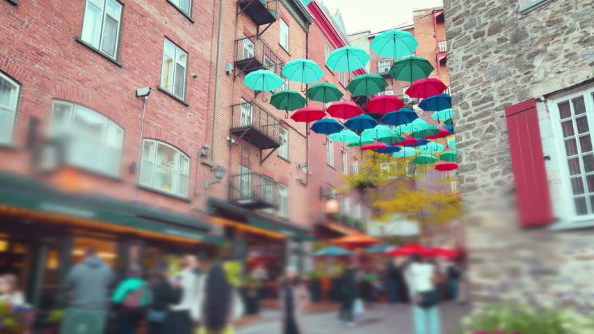 Umbrella Alley on Rue du Cul-de-Sac in Quebec City. Colorful umbrellas hang beautifully above the narrow historic street of Quartier Petit Champlain. Diverse crowd of tourists walks under the artistic canopy and visits local cafes. This lively autumn atmosphere showcases the popular Canadian landmark. Out of focus.
