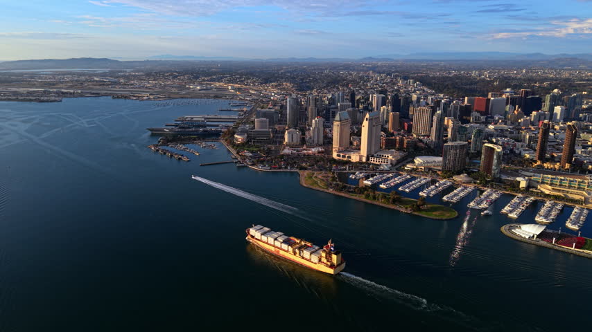 Container ship in San Diego Bay beside downtown marina, California. Cargo containers on deck, wake trail, yacht berths, pier, convention center sails, warship and tall hotels.