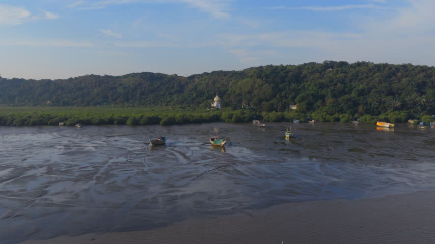 Drone flies from low tide mudflats and fishing boats over mangroves to reveal a white domed mausoleum on a green hillside in Mumbai, India.