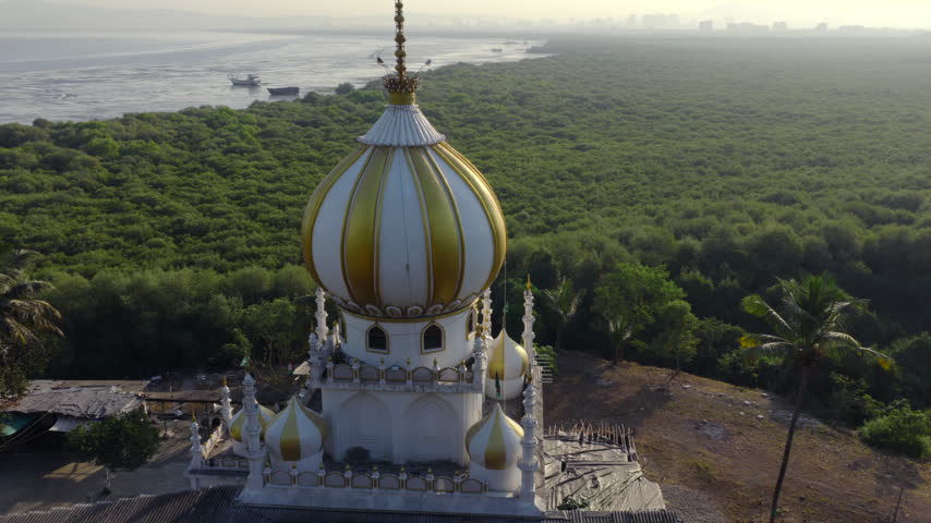 Drone push in and tilt up toward a white and gold domed mausoleum above mangroves with the sea behind in Mumbai, India.