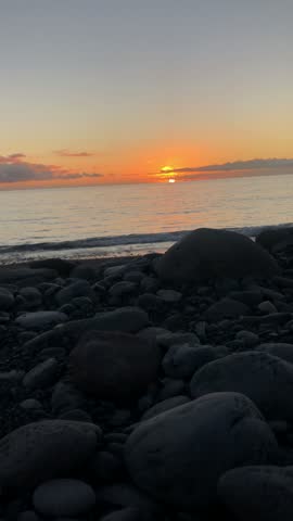 Vertical video of the sun setting over the ocean seen from a beach of volcanic pebbles on the coast of Tenerife, Spain