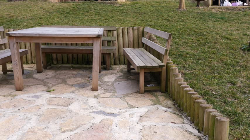 Slow pan view showing empty outdoor dining area featuring sturdy wooden table with benches on stone paved ground surrounded by green grass lawn and log border in recreational park.