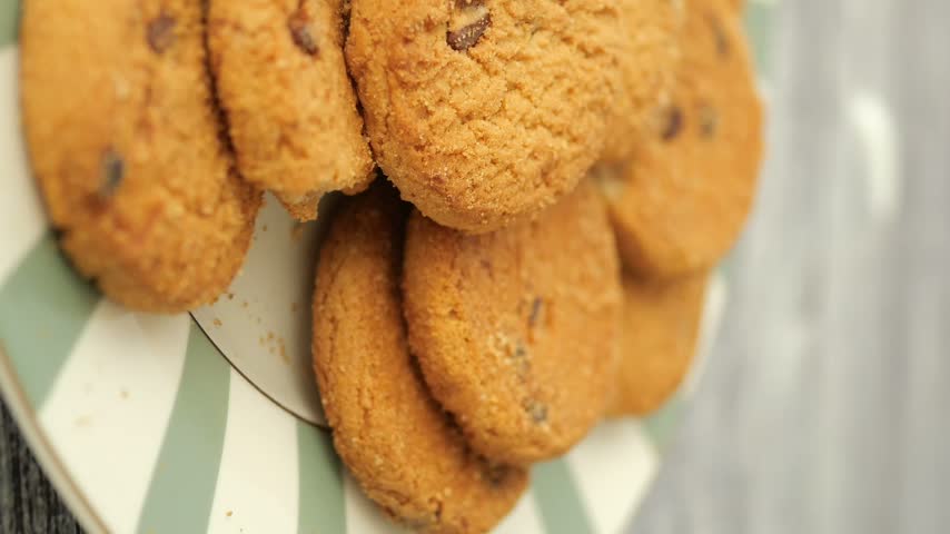 Close up cinematic shot of stacked chocolate chip cookies resting on a patterned ceramic plate featuring soft natural lighting ideal for food industry or culinary projects.
