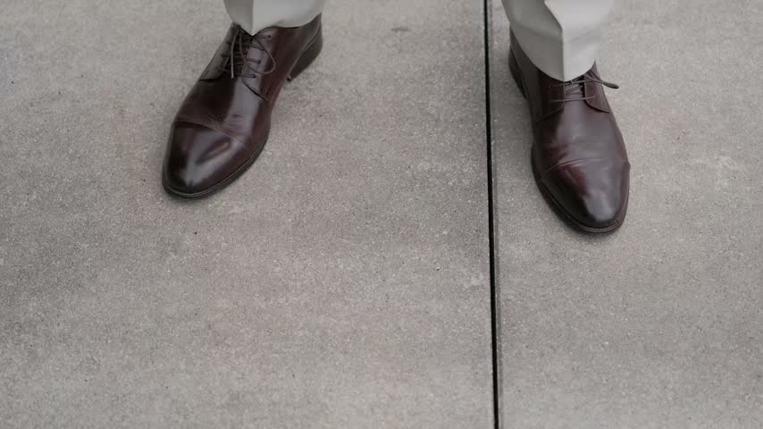 Close up of a man in brown leather shoes and beige trousers walking on concrete pavement