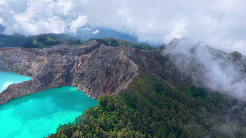 Aerial view of a turquoise crater lake and rugged volcanic ridge surrounded by clouds and forest at Mount Kelimutu, Flores, Indonesia.