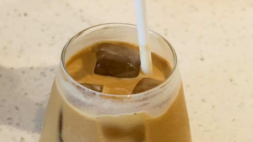 Cold brew iced coffee stirred with paper straw in clear glass on light marble table top macro shot.