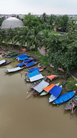 High angle view of traditional wooden boats with colorful tarps moored along the muddy riverbank in a tropical setting with lush green palm trees and a domed building in the background, Hoi An, Vietnam.