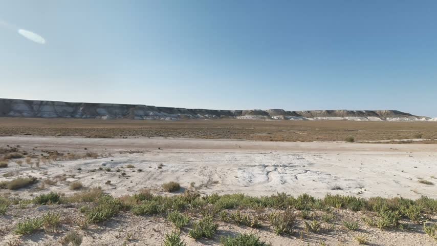 A moving roadside view passes scattered shrubs and pale rocky ground beneath a long, flat-topped escarpment across the open plain of Mangystau, Kazakhstan.