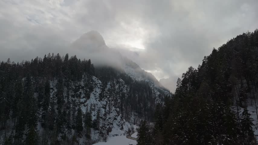 Drone view of a snow-covered forested gorge and winding river beneath misty rugged peaks in the Julian Alps near Kranjska Gora, Slovenia, in winter.