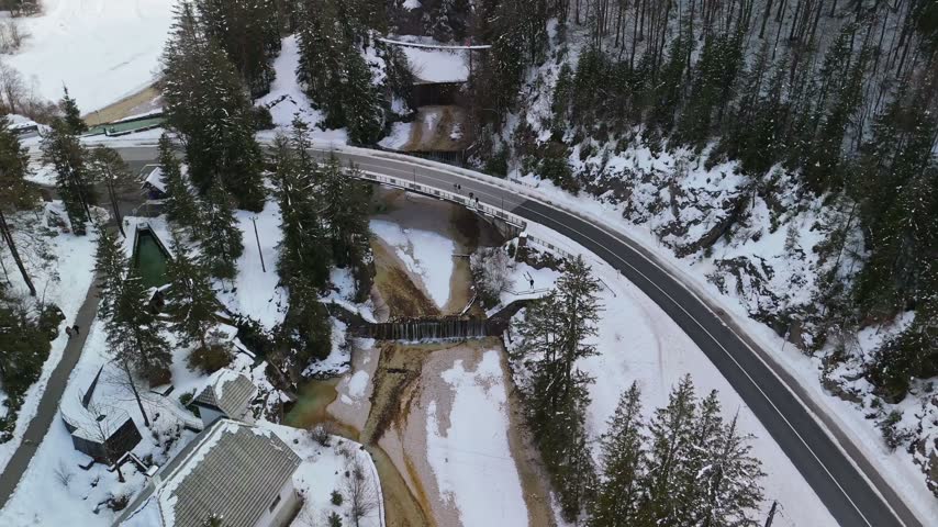 Aerial view of a road bridge above the Pišnica stream and waterfall within a steep snowy gorge amid conifer forest in Kranjska Gora, Slovenia, in winter.