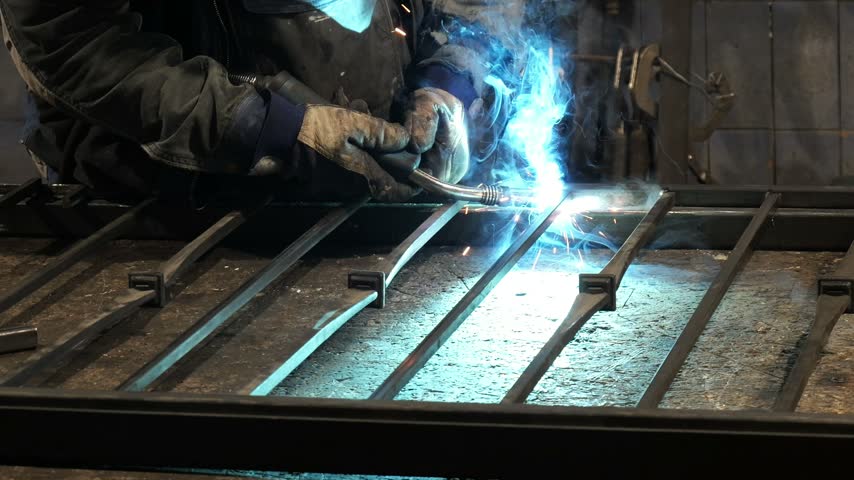 Professional welder joining steel parts of a metal gate with bright sparks flying in a workshop. A skilled blacksmith forging a wrought iron fence with an electric arc welding machine