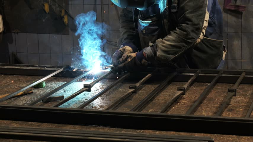 Metalworker wearing protective gear using an arc welding machine to assemble a steel gate, with bright blue light, hot sparks, and smoke rising from the molten metal in a dimly lit workshop