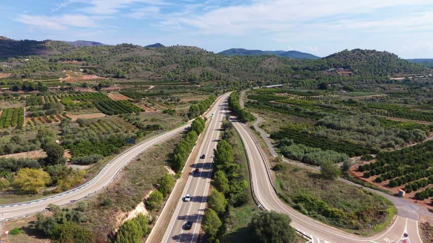 Multi-lane roundabout interchange cutting through citrus orchards and pine-covered hills under a partly cloudy sky, with light traffic on the highway