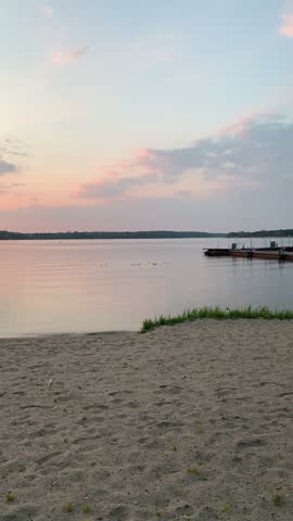 Slow pan along a sandy lake beach toward the orange setting sun, its reflection on calm water and two white swans at the shoreline.