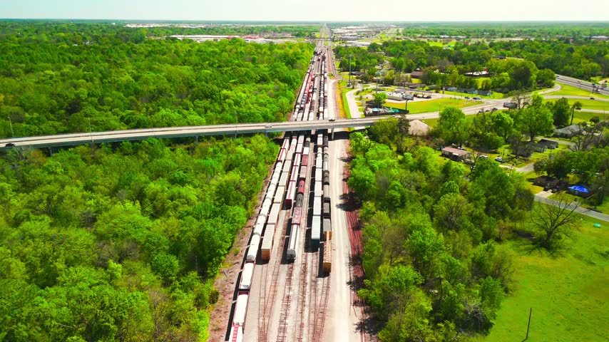 Aerial drone view along a Monroe Louisiana rail yard with freight cars beneath a road overpass, woodland and residential neighborhoods stretching toward the horizon