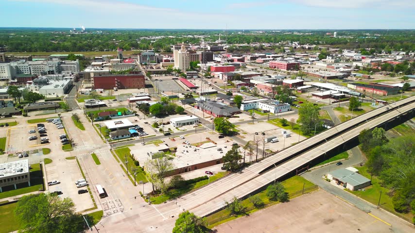 Aerial drone view of downtown Monroe, Louisiana, showing multistory buildings, intersecting streets, parking lots and an elevated highway amid green trees
