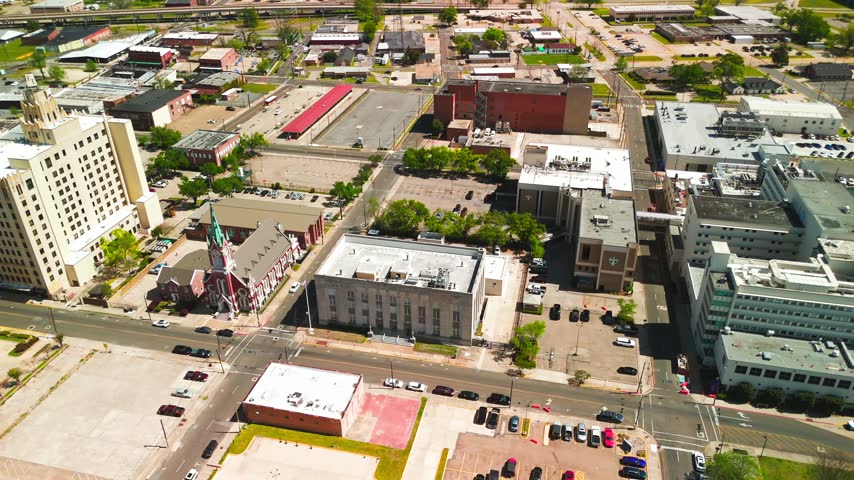 Oblique aerial drone view of downtown Monroe Louisiana with a tall cream tower, red brick church and green steeple above streets and parking lots