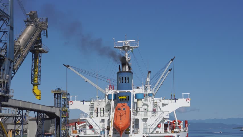 Large cargo ship, with dark exhaust smoke emitting from its funnel, is being unloaded at a pier next to industrial port machinery