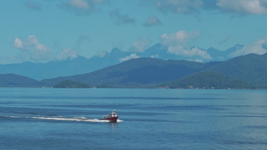 Small red and white pilot boat speeding across calm ocean waters, leaving a white wake with mountain ridges in the distance
