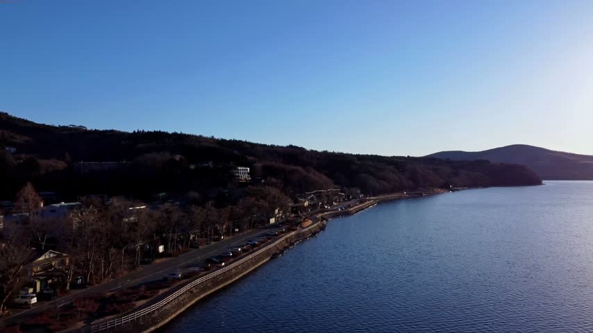 A curving shoreline road follows a blue lake below wooded hills, bare trees and scattered buildings under a clear sky.