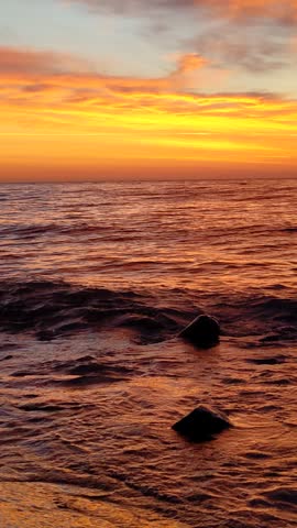 A vibrant sunrise paints the ocean sky with orange and yellow hues above gentle waves breaking on rocks.