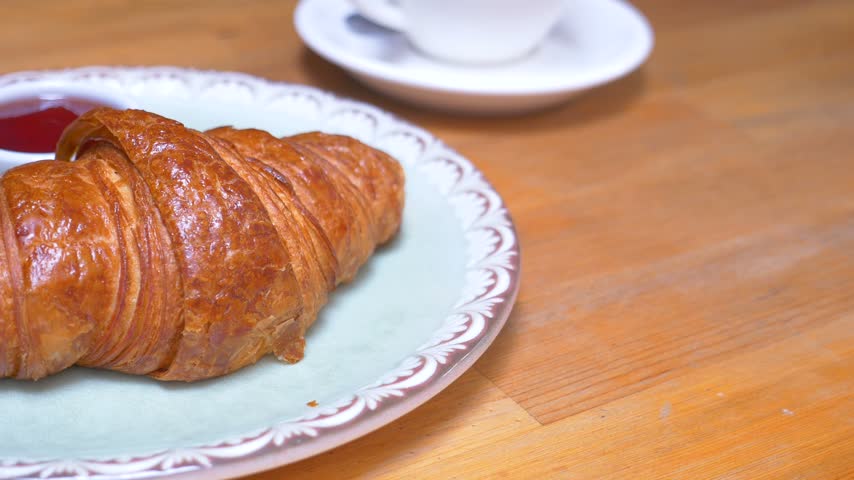 Appetizing golden buttery croissant served with small bowl of bright red jam on decorative plate placed on rustic wooden surface close up top view motion capture food footage.