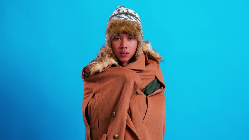 Freezing young man wrapped in thick coat and winter hat shivering from intense cold against blue background