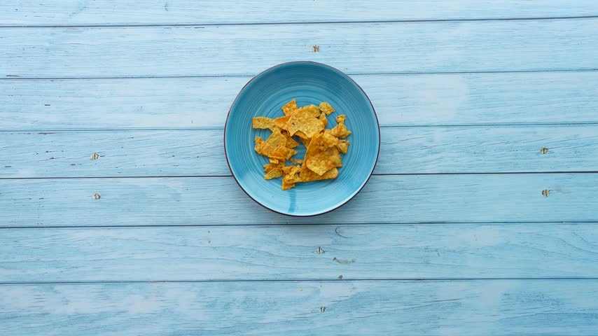 Person hands pouring golden corn tortilla chips from open plastic bag onto circular blue plate positioned on weathered light blue wood table surface high angle view slow motion clip.