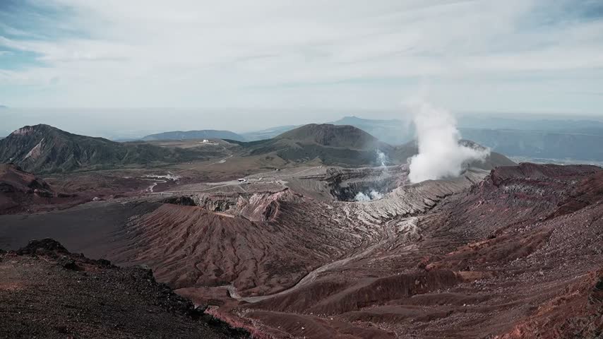 Aerial view of an the two Aso craters, an active volcano venting white steam amid rugged red and gray mountain terrain formin a Martian landscape