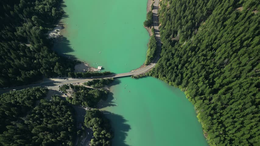 Top-down aerial drone shot showing a bridge and mountain road crossing the turquoise glacier water of Diablo Lake in North Cascades National Park. Kayaks float below on the vivid emerald water.