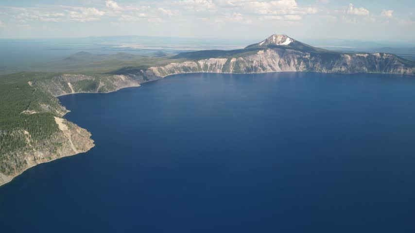 Aerial cinematic footage of Crater Lake National Park in Oregon, showcasing the vast caldera lake formed within an ancient volcano. The shot reveals the deep blue water surrounded by steep cliffs