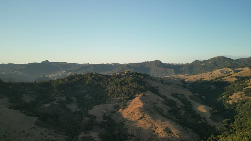 Aerial cinematic drone shot of Hearst Castle perched on the mountain ridge in San Simeon, California. Captured during golden hour, the castle and surrounding hills are bathed in warm sunlight.