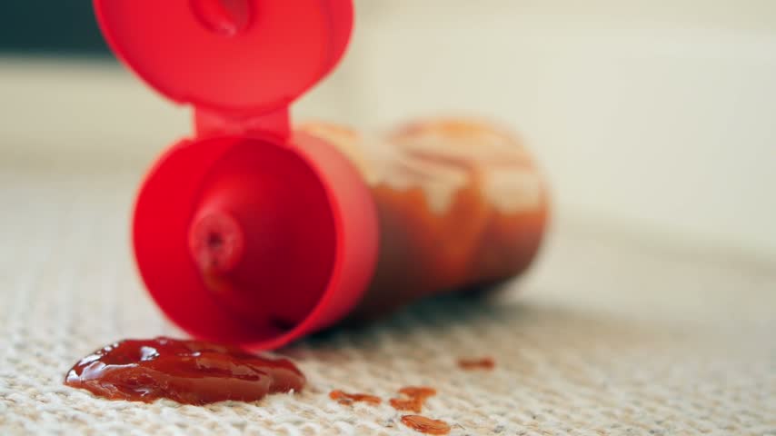 Close up video clip capturing spilled red tomato ketchup sauce on beige carpet with plastic container lying open nearby. Household accident scene showing messy kitchen or dining area floor.